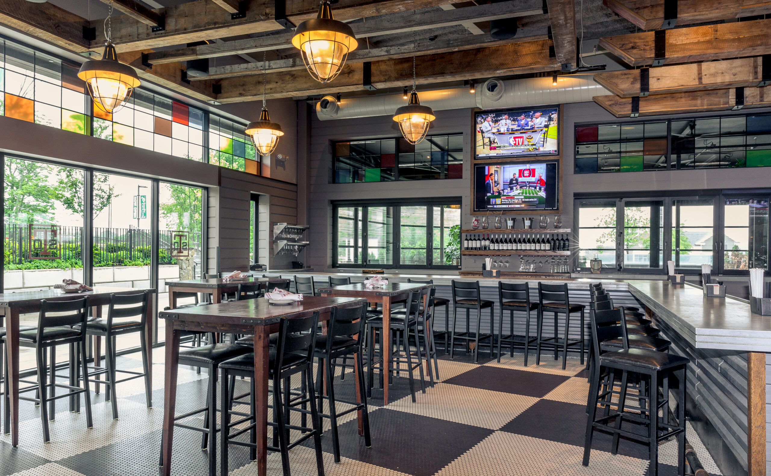 Modern sports bar interior in Littleton MA with high tables, bar seating, large windows, overhead lights, and two TVs above the bar showing sports channels.