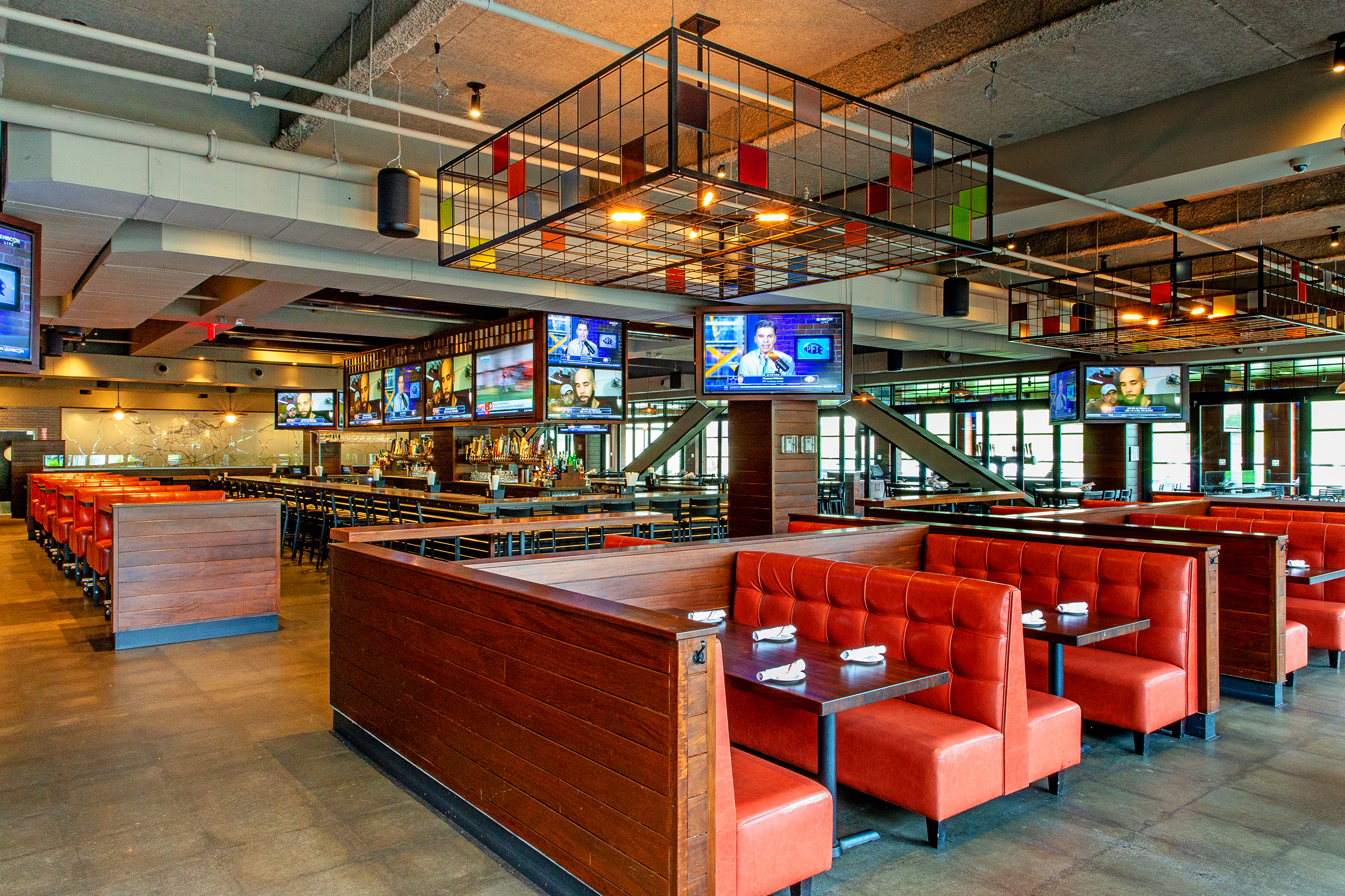 Spacious restaurant interior in Lowell MA with red booth seating, wooden tables set with napkins, multiple TVs on the walls, and modern industrial lighting fixtures.