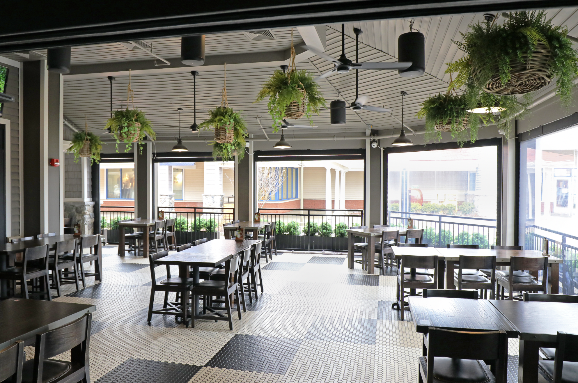 A covered outdoor patio area in Wrentham MA with dark wooden tables and chairs, hanging potted plants, tiled flooring, and large open windows letting in natural light.