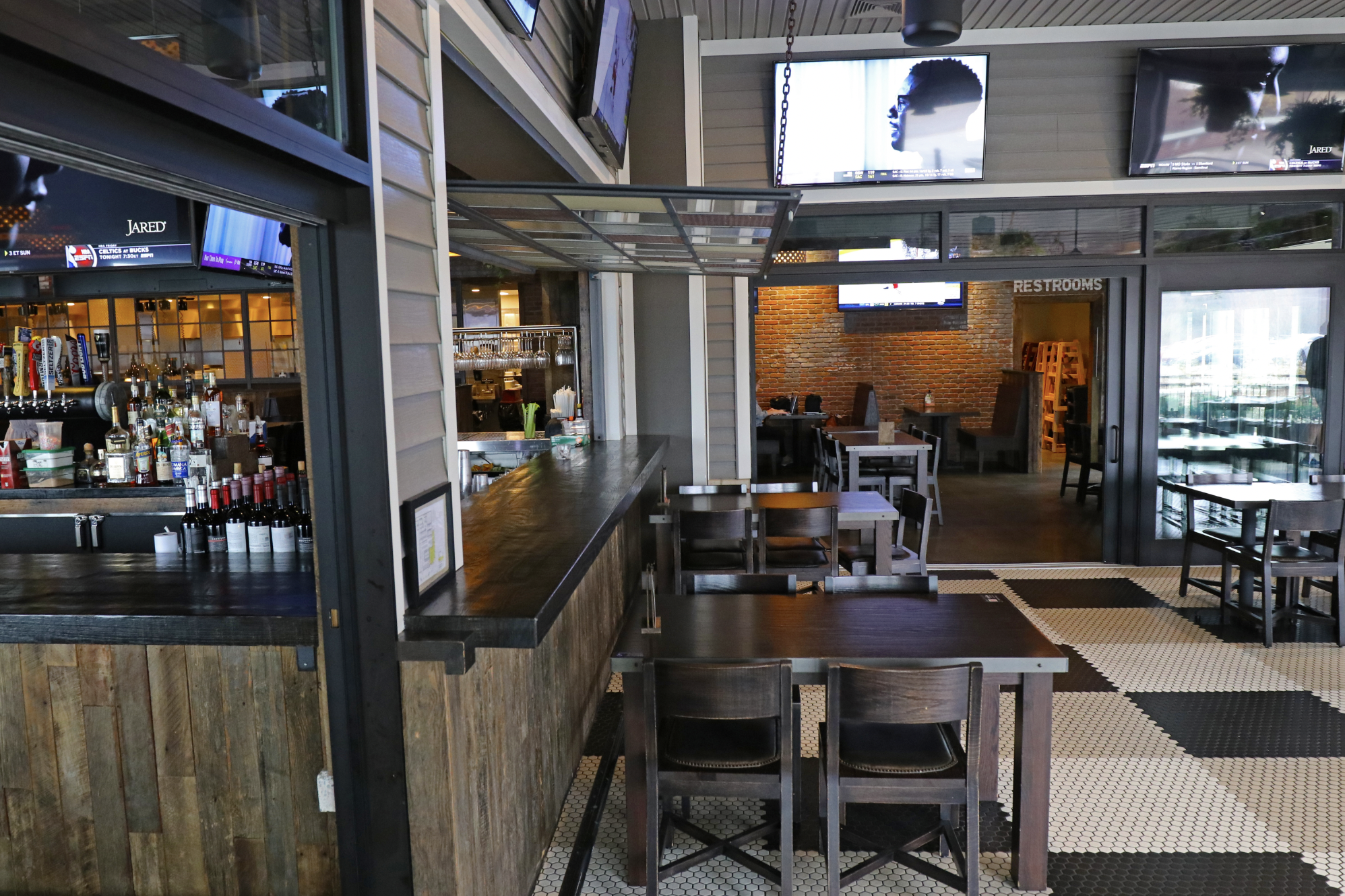 A modern bar and dining area in Wrentham MA, featuring wooden tables, a sleek bar counter, visible liquor bottles, and TV screens on the walls. A sign clearly points toward the restrooms.