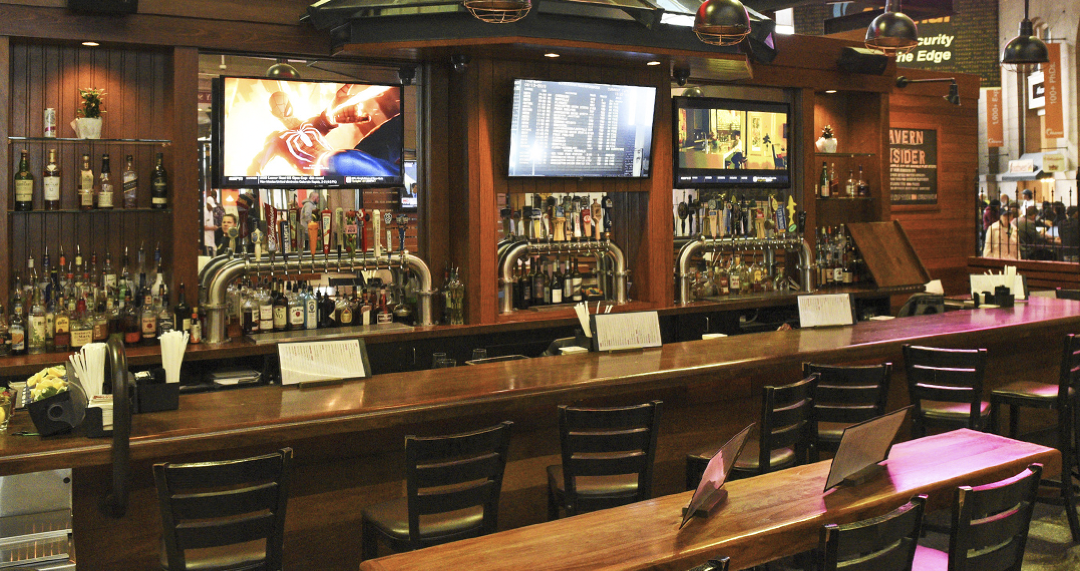 A wooden bar with empty chairs, numerous liquor bottles, taps, and three large TVs displaying various programs above the counter—just steps from South Station MA.