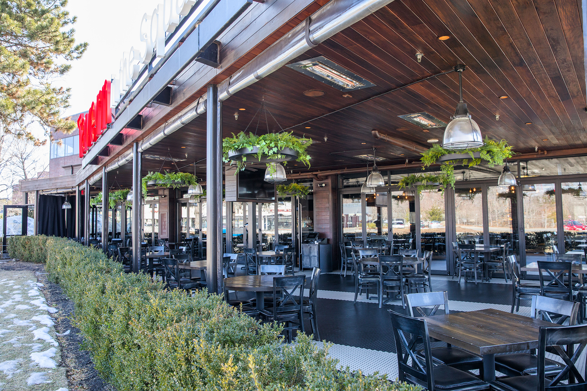 Outdoor restaurant patio in Burlington MA with wooden tables and black chairs, a partially covered roof, hanging plants, and surrounding greenery. A few patches of snow are visible on the ground outside.