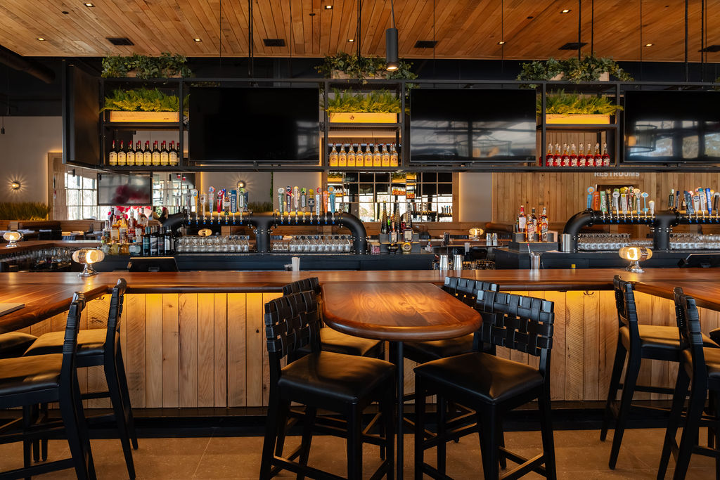 Modern bar interior with wooden accents, high tables and chairs, multiple beer taps, and flat-screen TVs above the counter. Shelves with bottles are visible in the background.