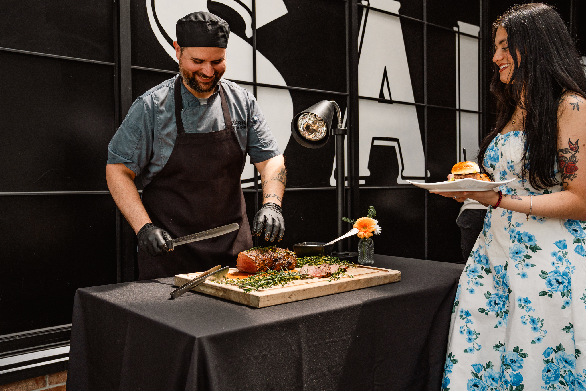 A chef slices roasted meat on a wooden board while a woman in a floral dress holds a plate with a sandwich, both standing by a black table—capturing the lively spirit often found at festive events.