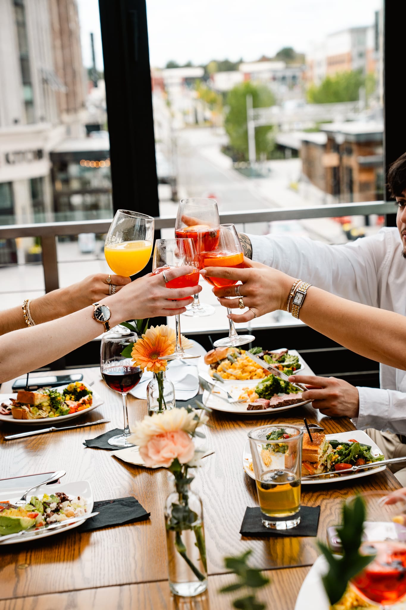 Four people clink glasses over a meal at a restaurant table set with plates of food, drinks, and flowers, celebrating special events against the backdrop of a stunning city view.