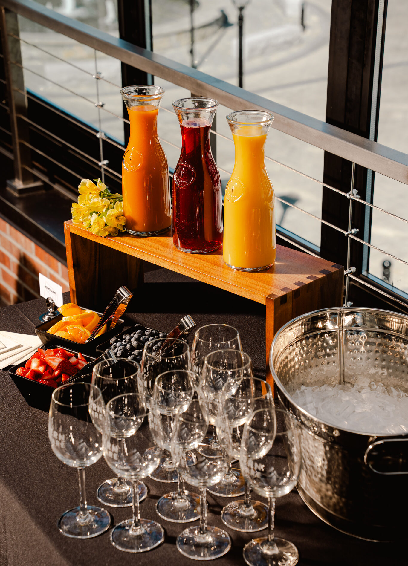 Three carafes of juice, a bowl of ice, assorted fruit, and rows of empty wine glasses are arranged on a black tablecloth near a window—perfectly set up for events or gatherings.