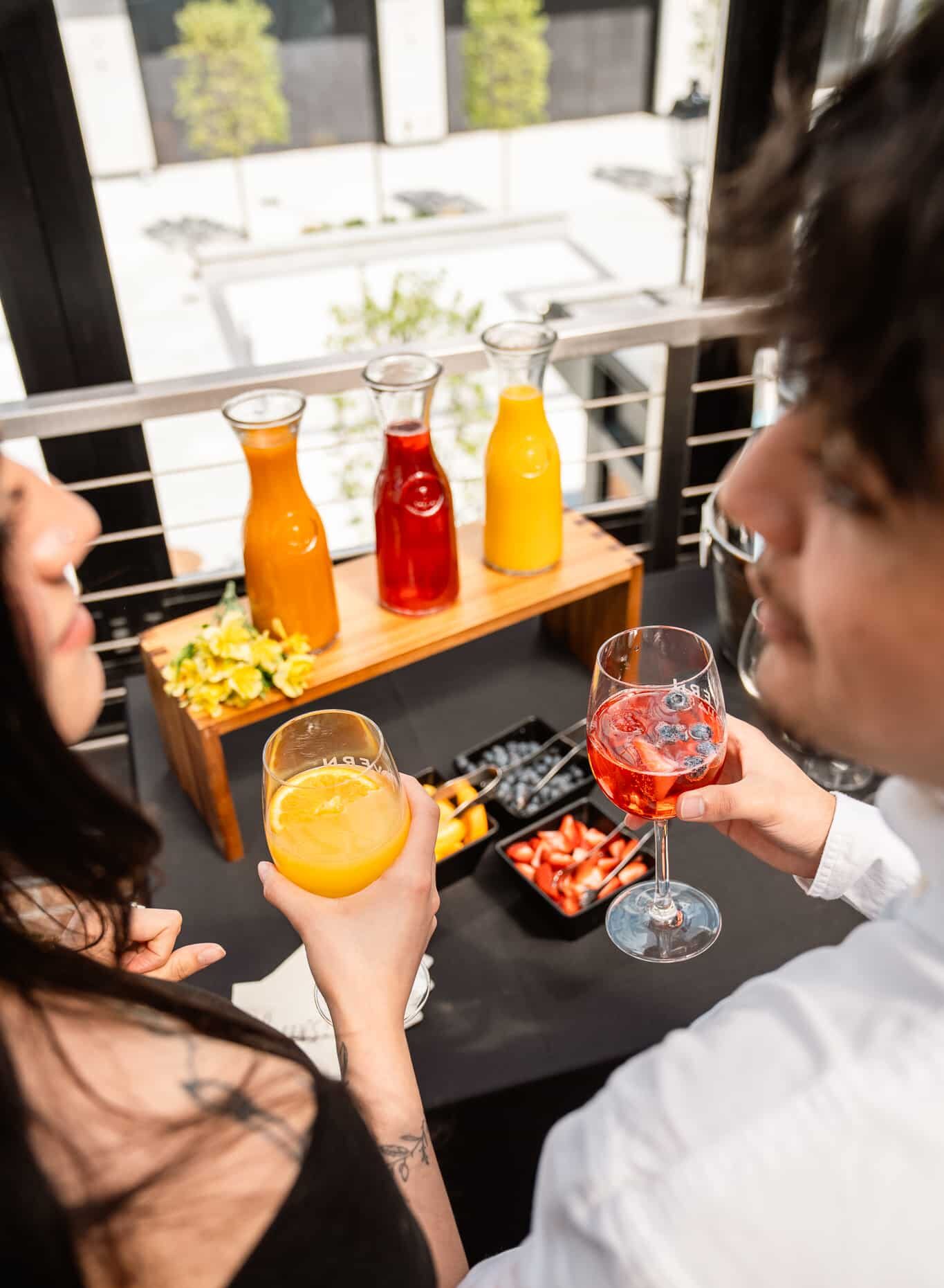 Two people holding colorful drinks stand by a table with juice carafes, fruit, and garnishes on an outdoor patio, creating the perfect setting for memorable events.