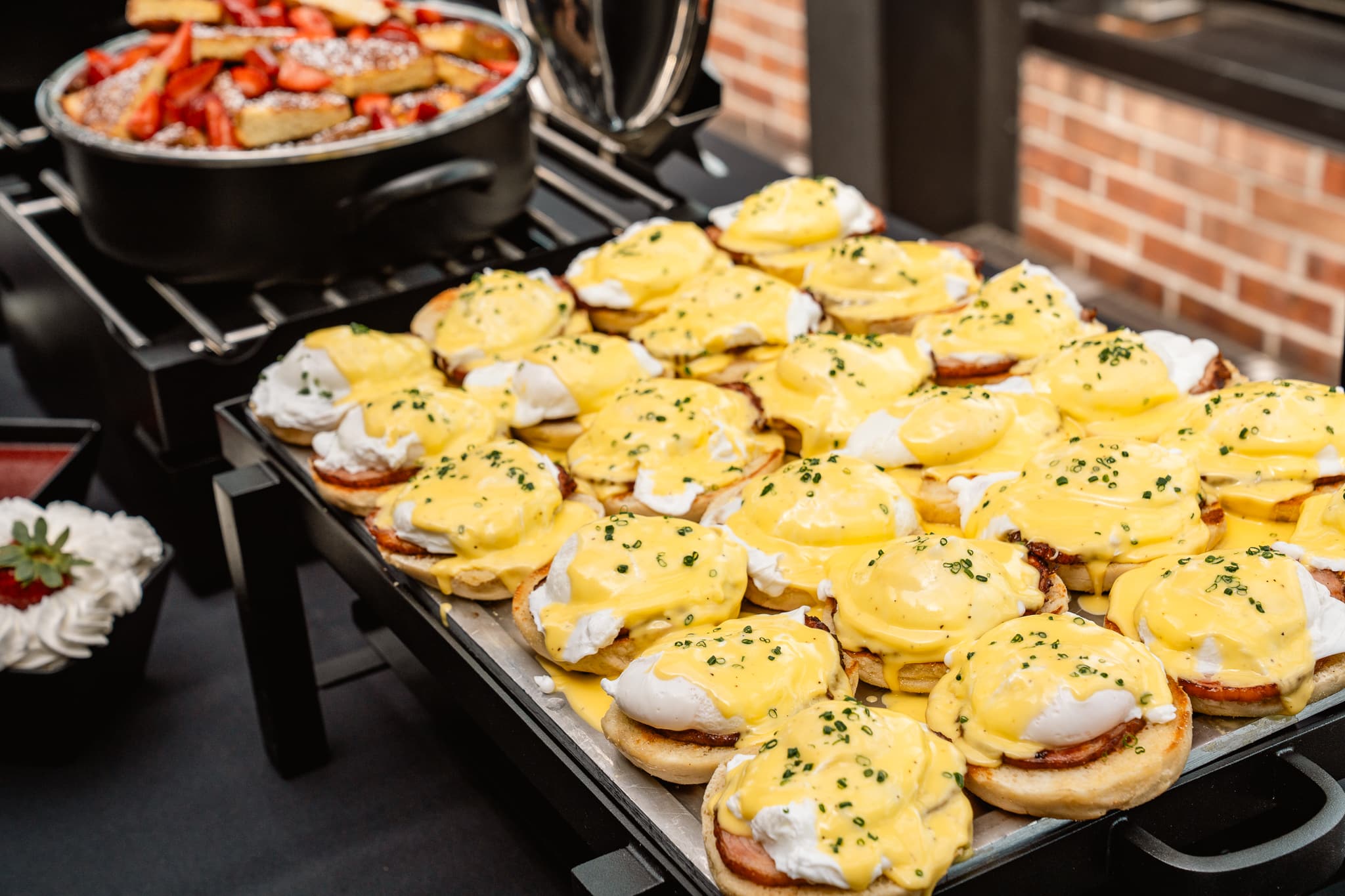 A tray of eggs Benedict topped with hollandaise sauce sits on a buffet table at events, next to a pan of roasted vegetables and a bowl of whipped cream with a strawberry garnish.