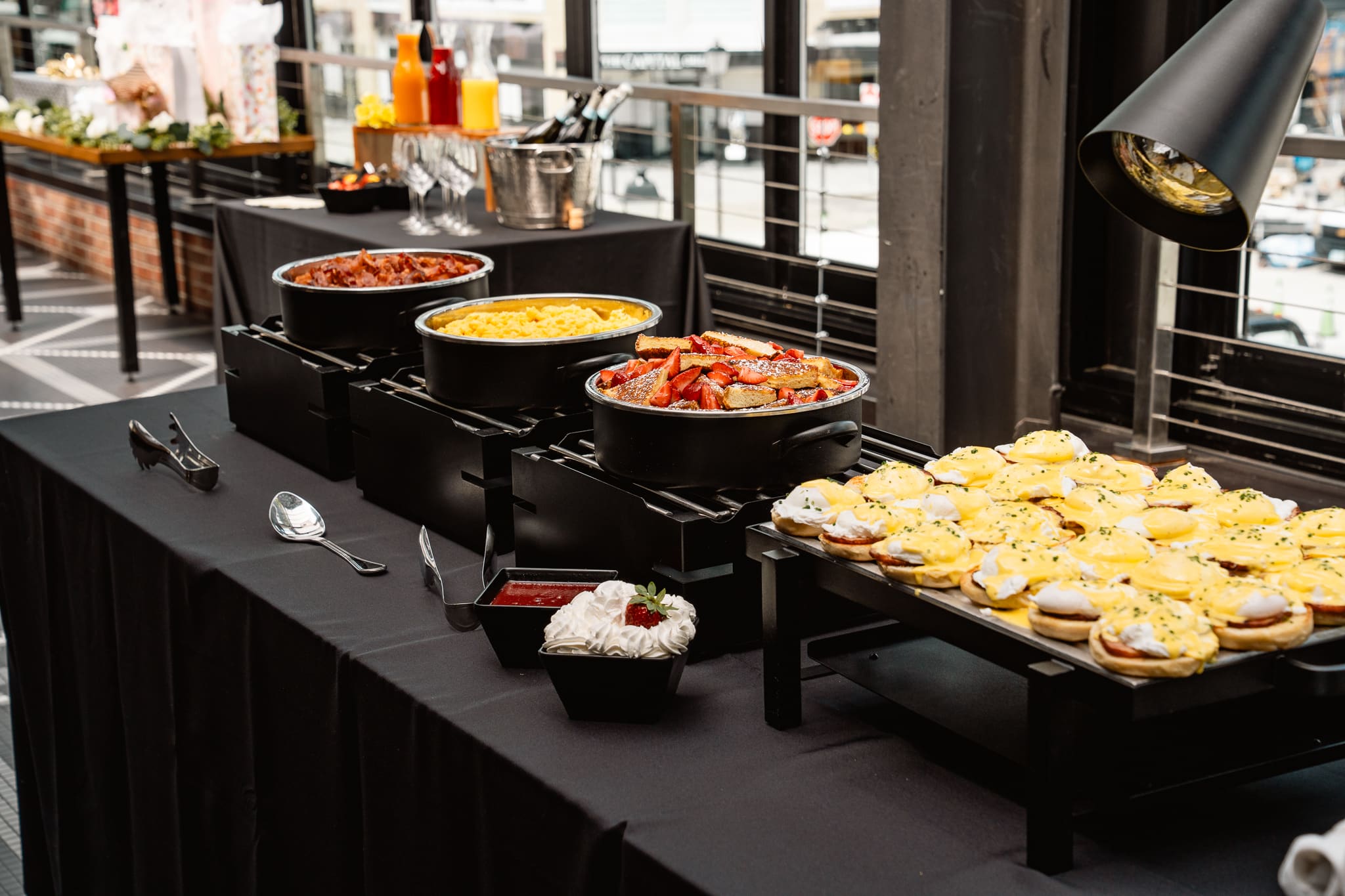 A buffet table is elegantly arranged for events, featuring chafing dishes filled with food, trays of eggs Benedict, a bowl of strawberries, and utensils set atop a black tablecloth in a bright room.