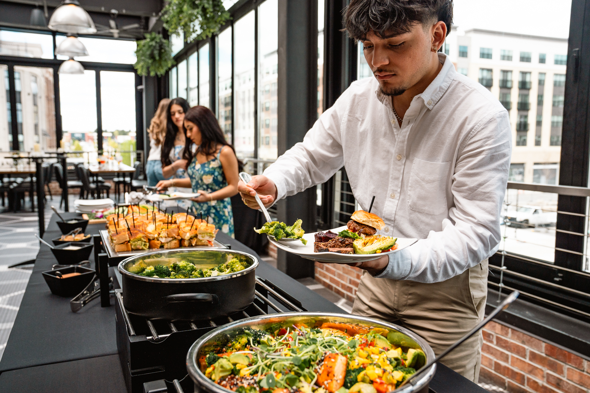 A man serves himself food from a buffet table with various salads and bread during an events gathering, while others wait in line behind him in a modern, sunlit dining area.