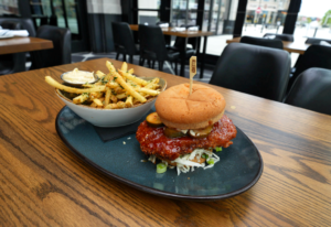 A breaded chicken sandwich with pickles and sauce sits on a plate next to a bowl of seasoned fries on a wooden table in a modern restaurant, making it a delicious choice for Winter Pairings.