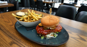 A breaded chicken sandwich with pickles and sauce sits on a plate next to a bowl of seasoned fries on a wooden table in a modern restaurant, making it a delicious choice for Winter Pairings.
