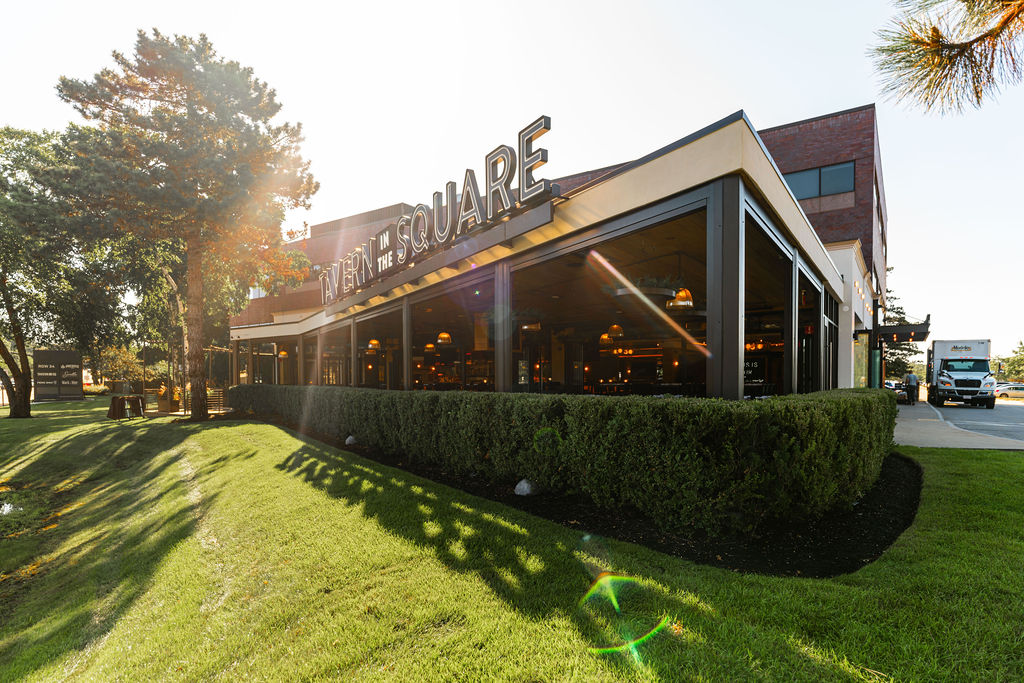 A modern restaurant named “TAVERN IN THE SQUARE” in Burlington MA, with large windows, surrounded by trimmed hedges and trees, on a sunny day.