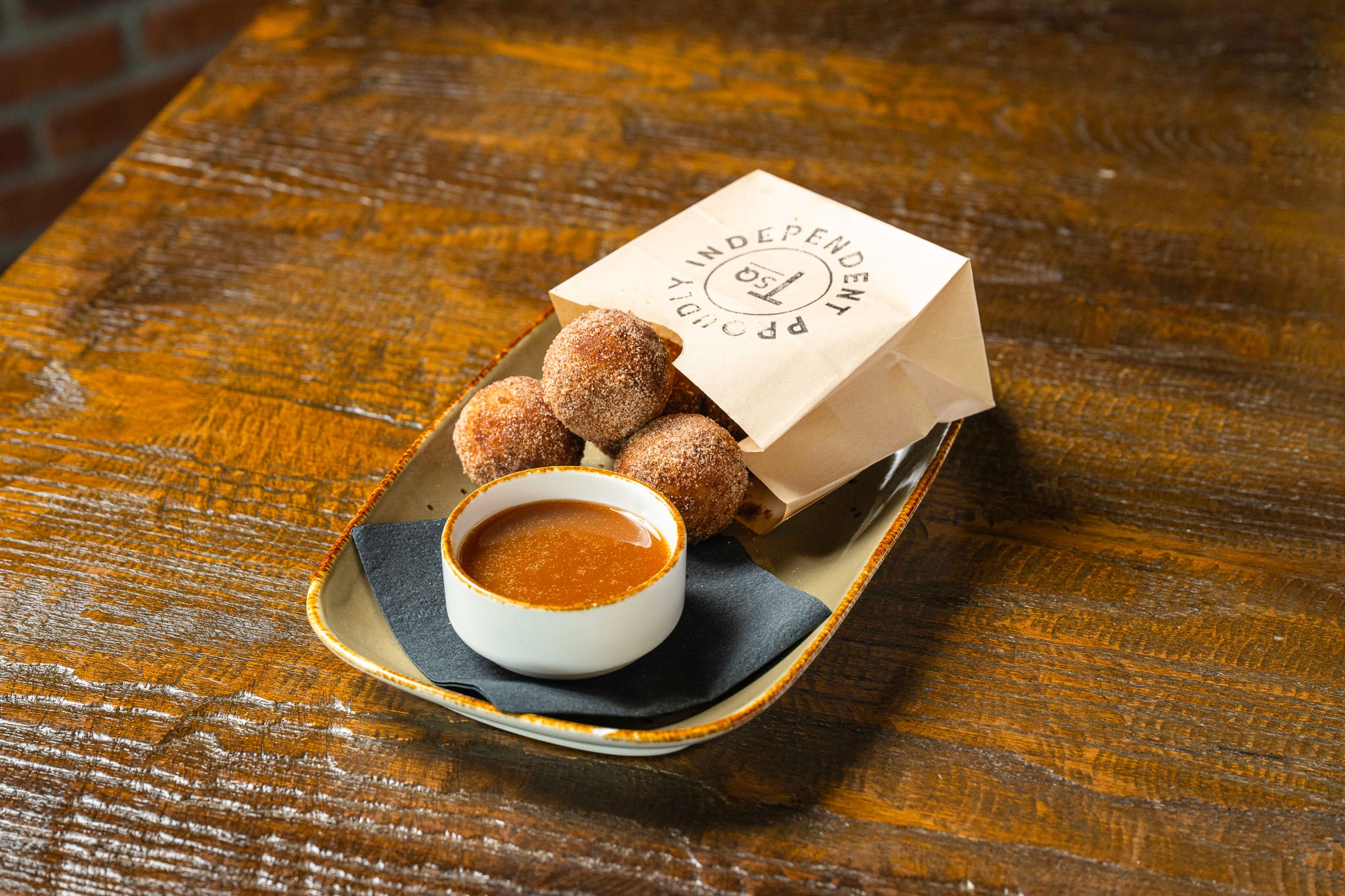 A plate with sugared donut holes beside a cup of caramel dipping sauce, served on a black napkin with a branded paper bag on a wooden table—perfect for enjoying at any of our Locations.